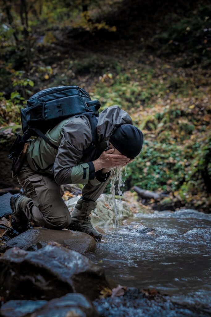 Man in outdoor gear drinking water from stream in forested area.